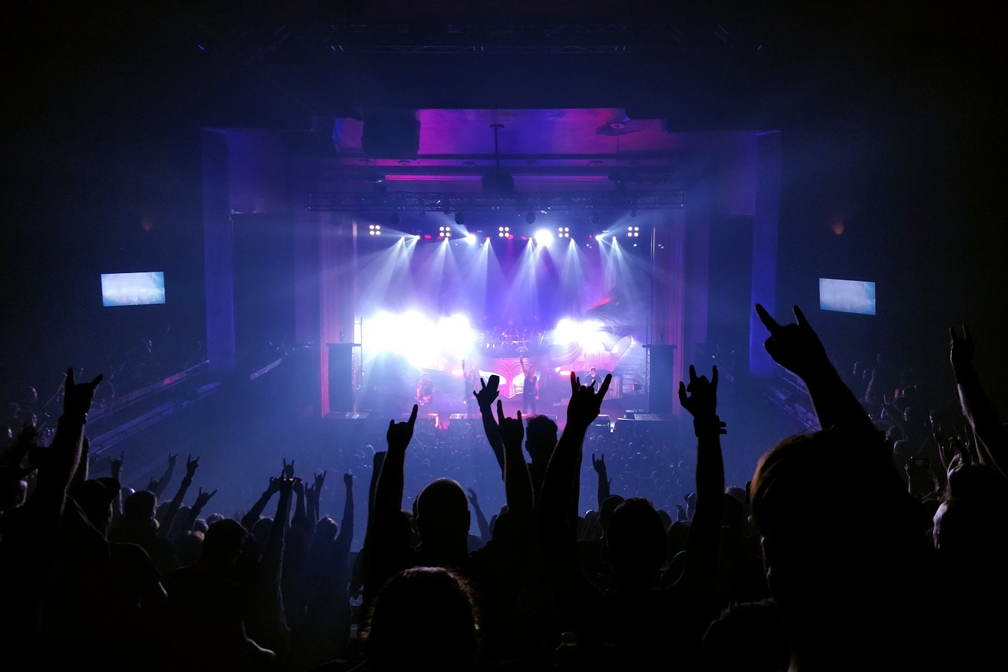 Concert audience with raised hands facing a brightly lit stage.